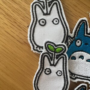 May include: Embroidered patches featuring a white creature with a sprout on its head, outlined in black. The sprout has green leaves. Other patches with blue and white characters are also visible.