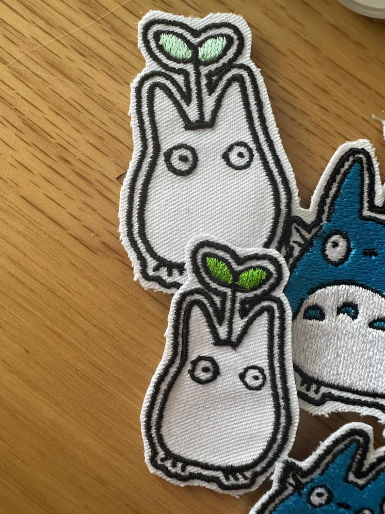 May include: Embroidered patches featuring a white creature with a sprout on its head, outlined in black. The sprout has green leaves. Other patches with blue and white characters are also visible.