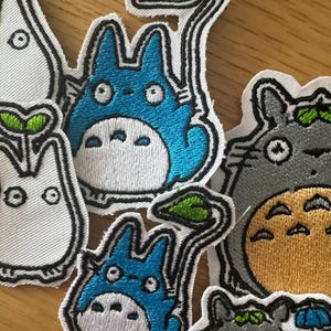 May include: Embroidered patches of cartoon characters in various colours. The patches include a blue character with a white belly and a green leaf, a grey character with a yellow belly, and a white character with a green leaf.
