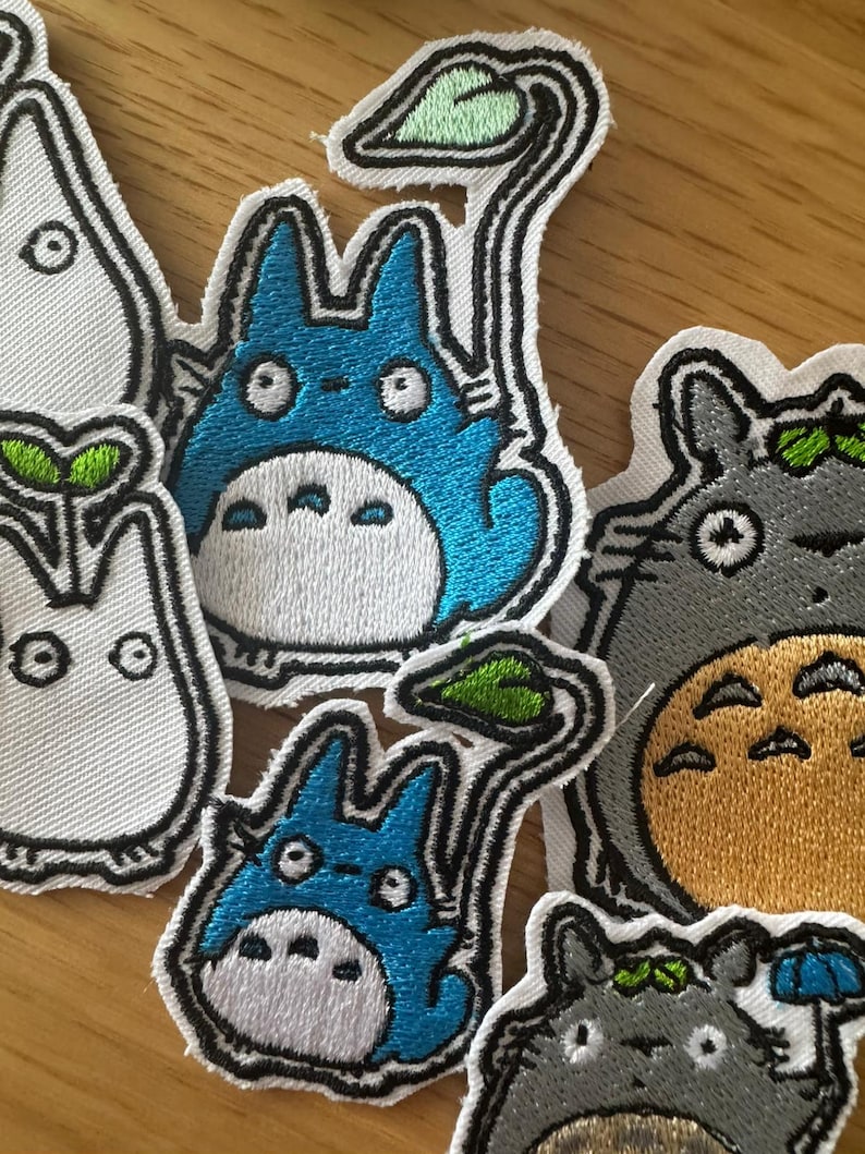 May include: Embroidered patches of cartoon characters in various colours. The patches include a blue character with a white belly and a green leaf, a grey character with a yellow belly, and a white character with a green leaf.