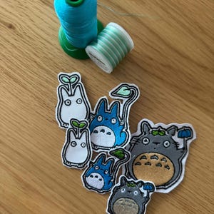 Può includere: Toppe ricamate con personaggi dei cartoni animati, tra cui Totoro, in varie dimensioni e pose. Due rocchetti di filo, uno turchese e uno a righe verdi e bianche, sono su una superficie di legno.