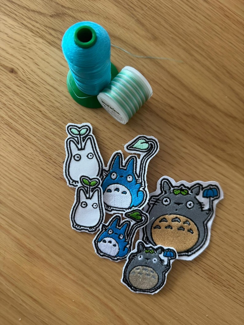 May include: Embroidered patches of cartoon characters, including Totoro, in different sizes and poses. Two spools of thread, one teal and one green and white striped, are arranged on a wooden surface.