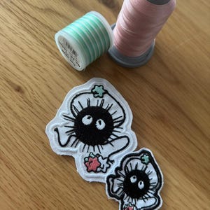 Può includere: Due toppe ricamate con un disegno di folletto di fuliggine nero, contorni bianchi e dettagli verdi e rosa. Sono visibili anche due rocchetti di filo, uno rosa e uno a righe verdi e bianche. Gli oggetti sono disposti su una superficie di legno.