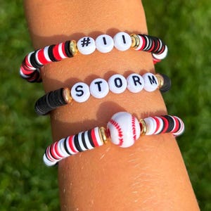May include: Three handmade beaded bracelets. The top bracelet has the text "#1" in white beads. The middle bracelet has the text "STORM". The bottom bracelet features a baseball bead. The bracelets have red, white, black, and gold accents.