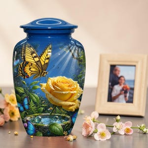 Könnte beinhalten: Blaue Urne mit Schmetterlings- und gelbem Rosen-Design. Die Urne zeigt eine detaillierte Illustration eines Schmetterlings, einer gelben Rose und eines kleinen Teiches mit Schmetterlingen. Ein gerahmtes Foto und Blumen sind im Hintergrund.