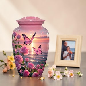 May include: A pink cremation urn with a lid, depicting a sunset scene with pink roses and butterflies. The urn has a glossy finish and a classic shape. A framed photo and flowers are in the background.