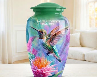 May include: A green-topped urn with a vibrant hummingbird and floral design. The urn features the name "JAMES ANDERSON" and the years "1965-2025". The hummingbird is depicted with colorful wings and a long beak, surrounded by pink and orange flowers.