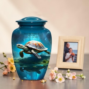 Puede incluir: Una urna de cremación turquesa con un diseño de tortuga y el texto "In Loving Memory of Alexa Pitt 2/4/1975-5/6/2025." La urna tiene una tapa a juego y un acabado brillante. Una foto enmarcada y flores están en el fondo.