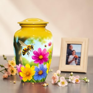May include: A yellow cremation urn with a bee and floral design. The urn features a bee hovering near colorful flowers, including pink, yellow, and blue blossoms. A framed photo and flowers are in the background.