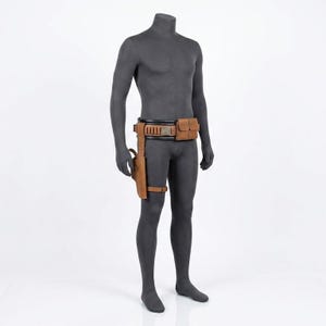 Puede incluir: Un cintur&oacute;n utilitario de cuero color canela sobre un maniqu&iacute; gris oscuro. El cintur&oacute;n presenta una banda interior negra, una bolsa rectangular, una serie de peque&ntilde;os bucles cil&iacute;ndricos y una funda de pierna sujeta con una correa al muslo.