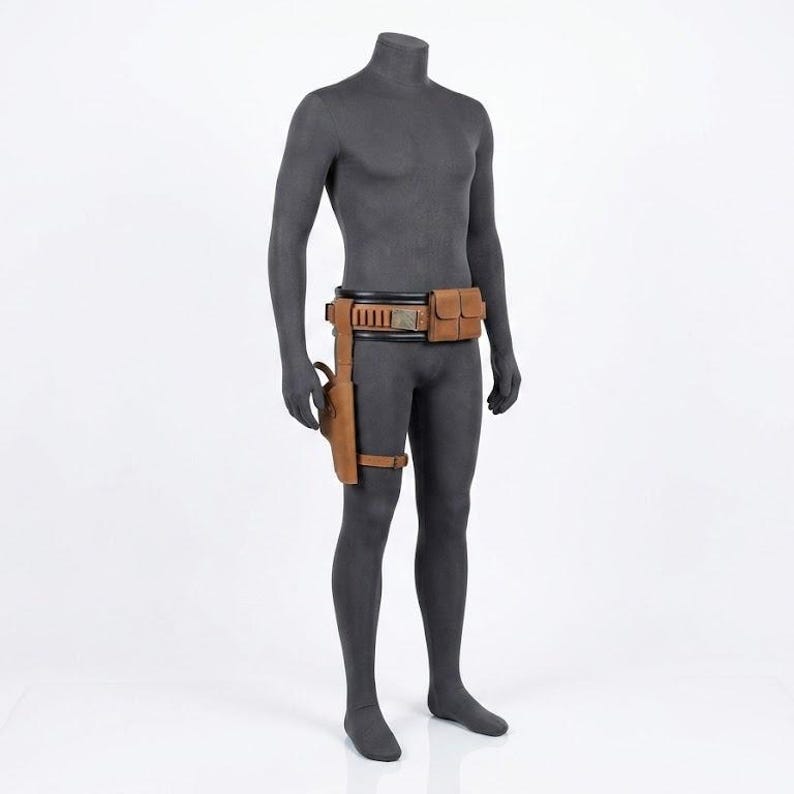Puede incluir: Un cintur&oacute;n utilitario de cuero color canela sobre un maniqu&iacute; gris oscuro. El cintur&oacute;n presenta una banda interior negra, una bolsa rectangular, una serie de peque&ntilde;os bucles cil&iacute;ndricos y una funda de pierna sujeta con una correa al muslo.