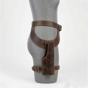 May include: A dark brown leather thigh holster featuring a waist belt and a leg strap. The holster is constructed with visible stitching and metal rivets, secured by metal buckles. The design includes a contoured hip piece and a vertical pouch.