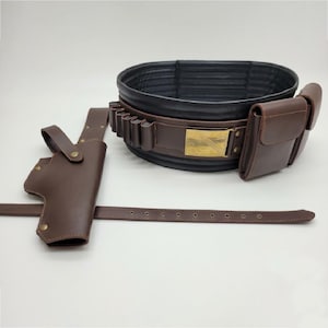 Puede incluir: Un cintur&oacute;n utilitario marr&oacute;n oscuro con forro interior negro, una hebilla rectangular color lat&oacute;n, m&uacute;ltiples trabillas integradas y bolsillos de almacenamiento. Una funda marr&oacute;n a juego con una correa larga se sit&uacute;a a la izquierda.