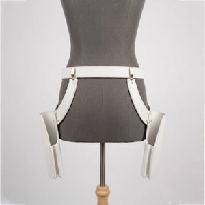 Op de afbeelding: Een wit dubbel holstergordelsysteem van leerachtig materiaal met een horizontale tailleband verbonden aan twee verticale zijholsters. Het ontwerp bevat zilverkleurige metalen hardware, klinknagels en clips die de holsters aan de riem bevestigen.
