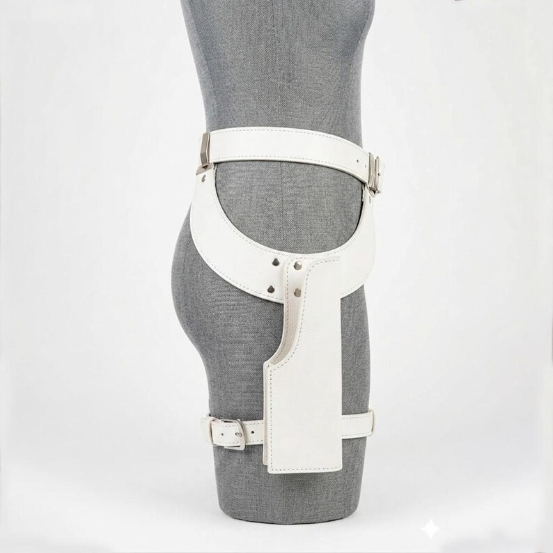 May include: A white leather thigh holster belt featuring a wide waist strap, a vertical holster pouch, and a lower leg strap. The accessory is secured with silver-toned metal buckles and rivets, designed to be worn around the waist and upper thigh.
