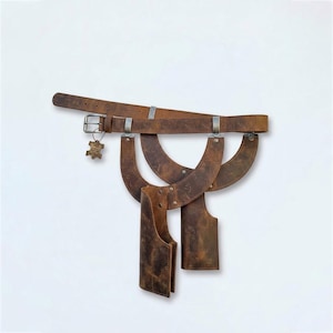 May include: A brown leather belt featuring a metal buckle and a small dangling tag. Attached to the belt are two curved leather panels and two vertical leather holsters, all secured with metal rivets and hardware against a plain white background.