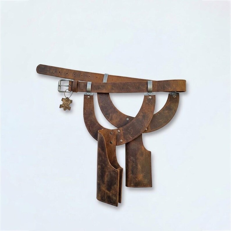 May include: A brown leather belt featuring a metal buckle and a small dangling tag. Attached to the belt are two curved leather panels and two vertical leather holsters, all secured with metal rivets and hardware against a plain white background.