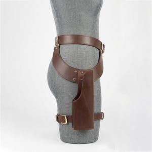 May include: A brown leather hip holster belt featuring a wide waist strap, a vertical holster pouch attached with silver-toned metal rivets, and a lower leg strap secured with a gold-toned metal buckle, worn on a gray mannequin torso.