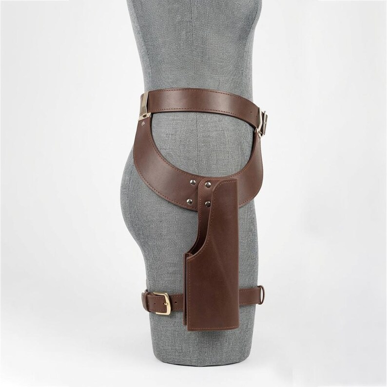 May include: A brown leather hip holster belt featuring a wide waist strap, a vertical holster pouch attached with silver-toned metal rivets, and a lower leg strap secured with a gold-toned metal buckle, worn on a gray mannequin torso.