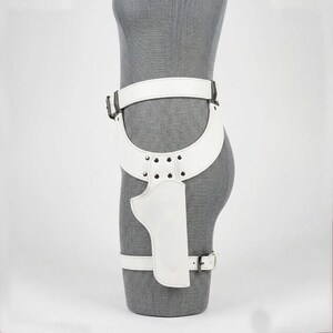 May include: A white leather thigh holster belt featuring a wide waist strap, a curved hip panel secured with silver-toned metal rivets, and a vertical holster pouch. The assembly includes an adjustable lower leg strap with a silver-toned metal buckle.