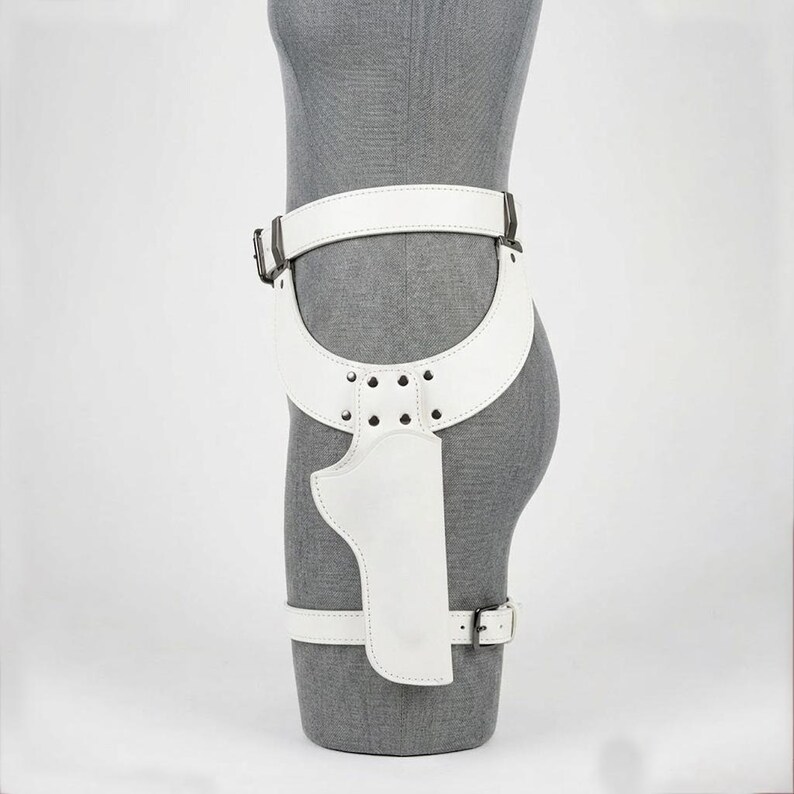 May include: A white leather thigh holster belt featuring a wide waist strap, a curved hip panel secured with silver-toned metal rivets, and a vertical holster pouch. The assembly includes an adjustable lower leg strap with a silver-toned metal buckle.