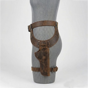 May include: A brown leather thigh holster featuring a wide waist belt and a secondary leg strap, both secured with metal buckles. The holster is constructed with riveted panels and displays a distressed, weathered texture throughout the surface.