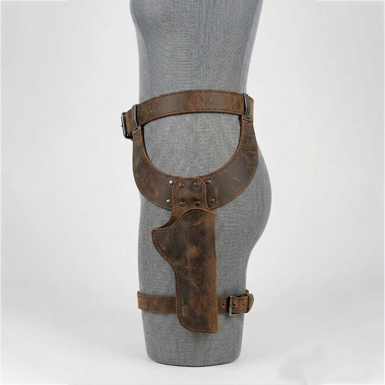 May include: A brown leather thigh holster featuring a wide waist belt and a secondary leg strap, both secured with metal buckles. The holster is constructed with riveted panels and displays a distressed, weathered texture throughout the surface.