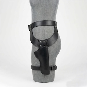 May include: A black leather thigh holster featuring a wide waist belt connected to a molded holster pouch. The design includes adjustable straps with metal buckles and silver-toned rivets securing the holster to the leg and waist area.