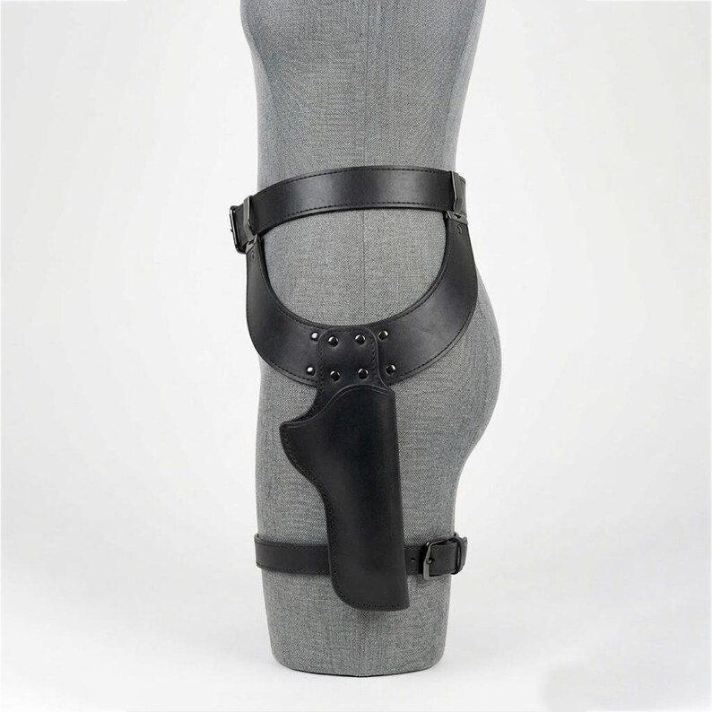 May include: A black leather thigh holster featuring a wide waist belt connected to a molded holster pouch. The design includes adjustable straps with metal buckles and silver-toned rivets securing the holster to the leg and waist area.