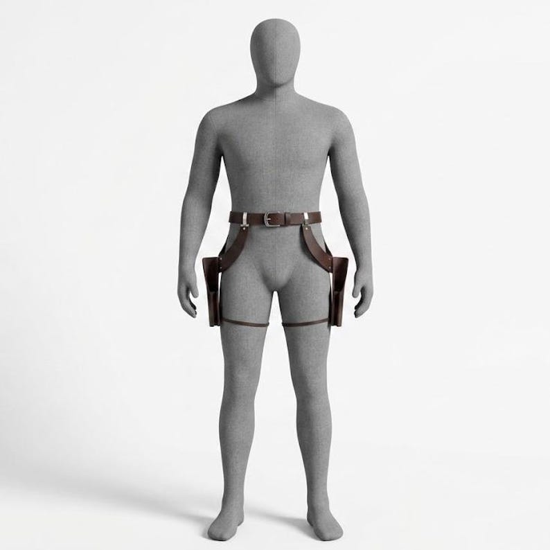 May include: A dark brown leather dual holster rig featuring a waist belt with a silver-toned metal buckle, two side-mounted holsters, and adjustable leg straps. The leather gear is displayed on a featureless gray mannequin against a white background.