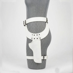 Op de afbeelding: Een witte lederen dijbeenholsterriem met een brede tailleband, een gebogen heuppaneel vastgezet met zilverkleurige metalen klinknagels en een verticale holsterzak. Het geheel bevat een verstelbare beenband met een zilverkleurige metalen gesp.