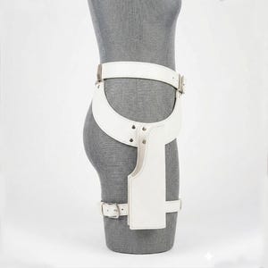Op de afbeelding: Een witte lederen dijbeenholstergordel met een brede heupband, een verticaal holsterzakje en een beenband. Het accessoire is bevestigd met zilverkleurige metalen gespen en klinknagels, ontworpen om rond de taille en het bovenbeen te worden gedragen.