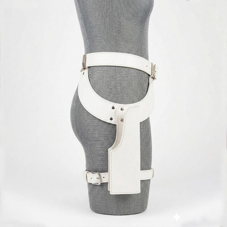 Op de afbeelding: Een witte lederen dijbeenholstergordel met een brede heupband, een verticaal holsterzakje en een beenband. Het accessoire is bevestigd met zilverkleurige metalen gespen en klinknagels, ontworpen om rond de taille en het bovenbeen te worden gedragen.