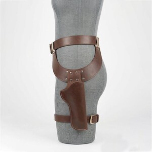 May include: A brown leather thigh holster featuring a wide waist belt and a secondary leg strap. The holster is constructed with silver-toned metal rivets and a rectangular buckle, designed to secure a sidearm against the upper leg area.