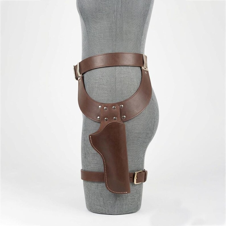 May include: A brown leather thigh holster featuring a wide waist belt and a secondary leg strap. The holster is constructed with silver-toned metal rivets and a rectangular buckle, designed to secure a sidearm against the upper leg area.