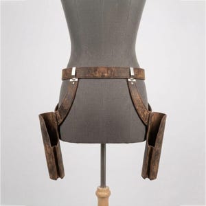May include: A brown distressed leather double holster belt featuring two vertical side pouches attached to a waist strap with silver-toned metal hardware. The holster system is displayed on a gray mannequin torso, showcasing a weathered texture.