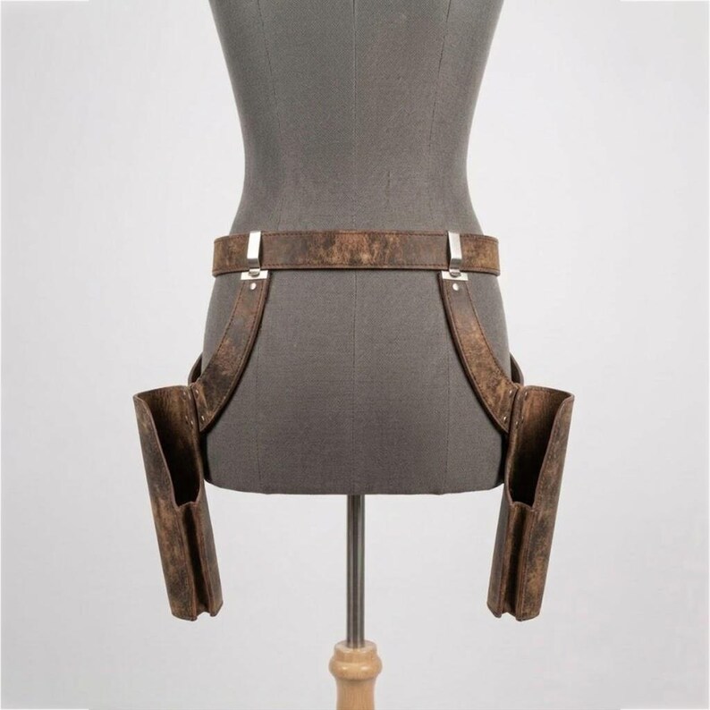 May include: A brown distressed leather double holster belt featuring two vertical side pouches attached to a waist strap with silver-toned metal hardware. The holster system is displayed on a gray mannequin torso, showcasing a weathered texture.