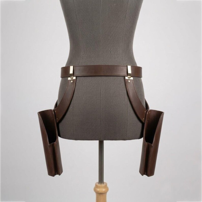 May include: A dark brown leather belt featuring two attached holster-style pouches hanging on either side. The belt is secured with metallic silver-toned hardware, including rectangular buckles and rivets connecting the pouches to the main waist strap.