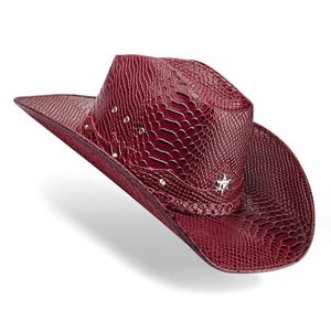 Może przedstawiać: Bordowy kapelusz kowbojski o fakturze skóry węża. Kapelusz posiada srebrne ćwieki i ozdobę w kształcie gwiazdy. Rondo jest zakrzywione, a kapelusz ma pleciony pasek wokół korony.