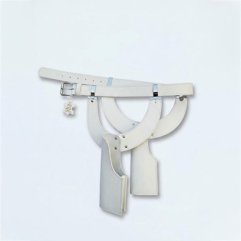 May include: A white leather belt featuring a silver-toned metal buckle and two attached holster-style pockets. The belt includes a small rectangular tag with text hanging from the buckle. The design consists of curved leather straps and silver rivets.