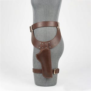 May include: A brown leather thigh holster featuring a wide waist belt and a secondary leg strap. The holster is constructed with reinforced silver-toned metal rivets at the connection point and includes a metal buckle on the lower leg strap.