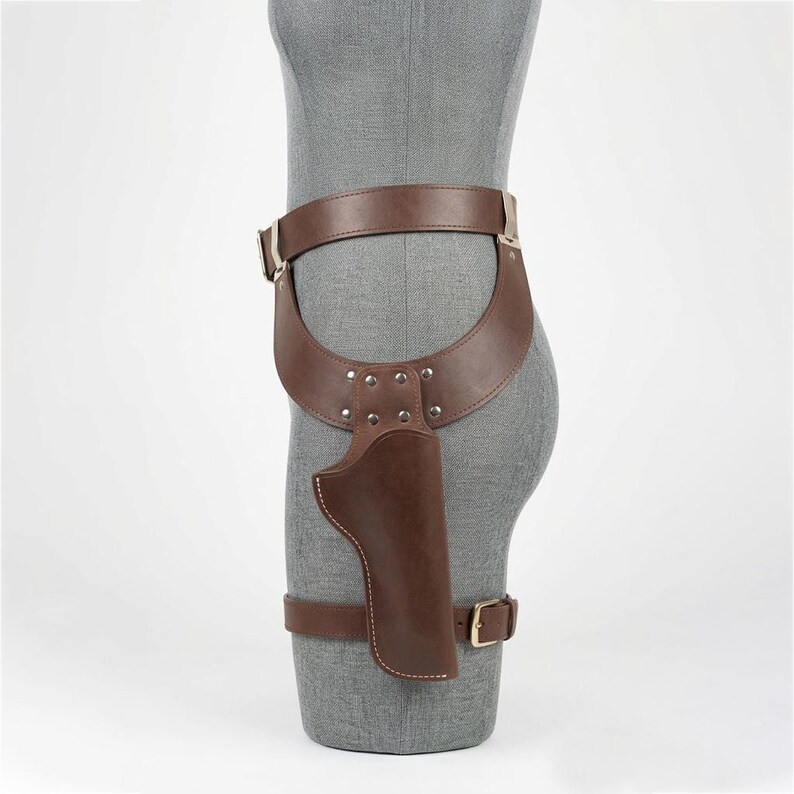 May include: A brown leather thigh holster featuring a wide waist belt and a secondary leg strap. The holster is constructed with reinforced silver-toned metal rivets at the connection point and includes a metal buckle on the lower leg strap.