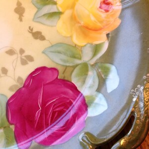 Puede incluir: Un plato de servir de porcelana vintage con un dise&ntilde;o floral. El plato presenta una rosa amarilla, una rosa rosa y hojas verdes. El plato tiene un borde dorado y un borde festoneado.