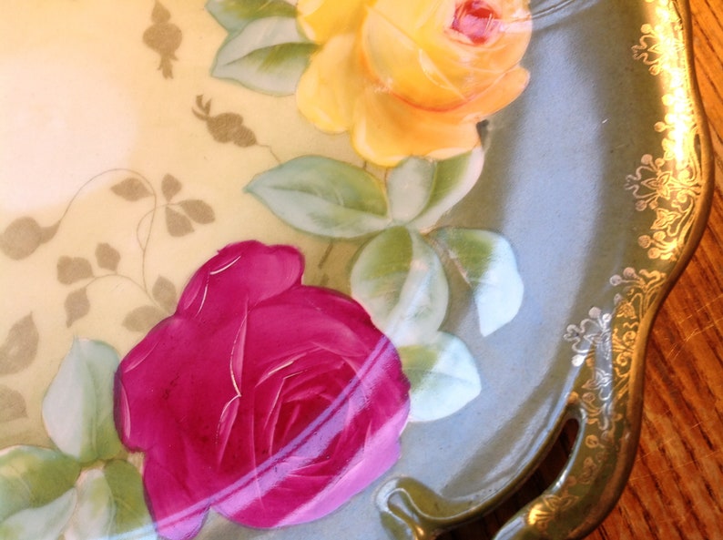 Puede incluir: Un plato de servir de porcelana vintage con un dise&ntilde;o floral. El plato presenta una rosa amarilla, una rosa rosa y hojas verdes. El plato tiene un borde dorado y un borde festoneado.