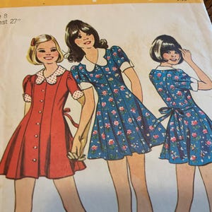 May include: Vintage sewing pattern featuring illustrations of three girls in dresses. The dresses include a red button-down dress with a white polka dot collar, and two blue floral dresses. The pattern is for size 8 with a 27-inch breast.