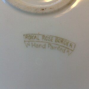 Puede incluir: Primer plano de un plato de cer&aacute;mica blanco con un sello dorado que dice "Royal Rose Border Hand Painted".