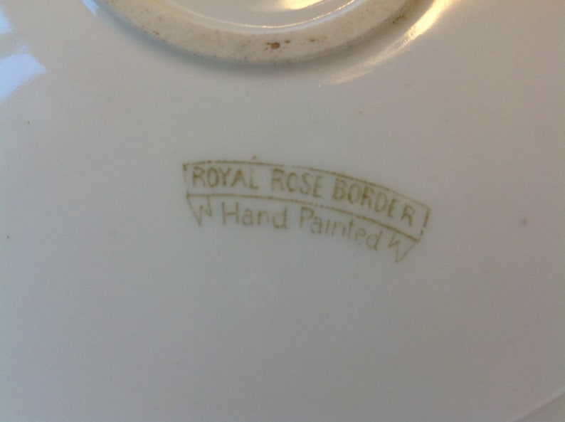 Puede incluir: Primer plano de un plato de cer&aacute;mica blanco con un sello dorado que dice "Royal Rose Border Hand Painted".
