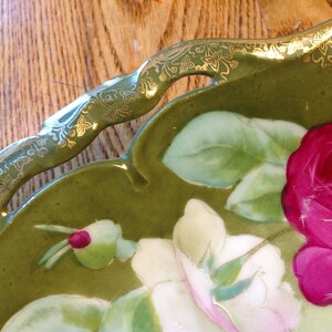 Puede incluir: Un plato de servir de cer&aacute;mica verde con un borde dorado y un dise&ntilde;o floral con rosas rosas y blancas. El plato tiene un borde festoneado y dos asas recortadas.