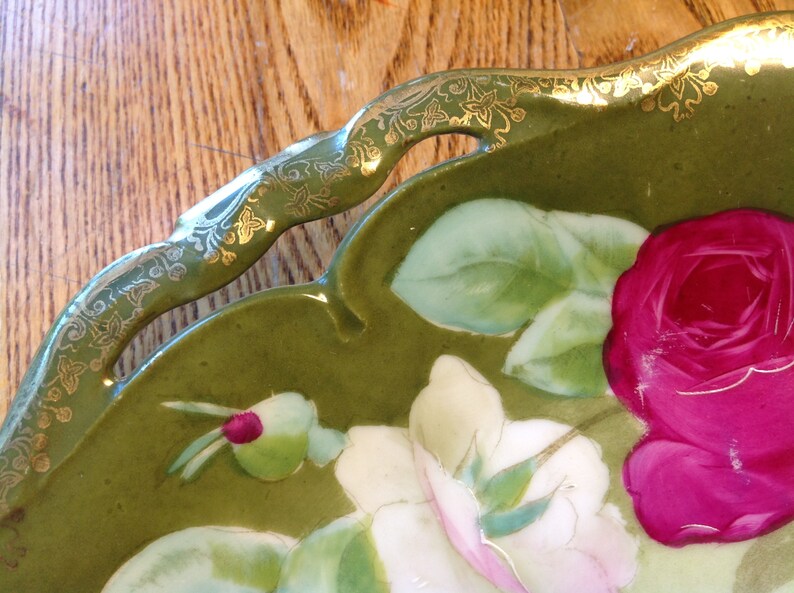 Puede incluir: Un plato de servir de cer&aacute;mica verde con un borde dorado y un dise&ntilde;o floral con rosas rosas y blancas. El plato tiene un borde festoneado y dos asas recortadas.