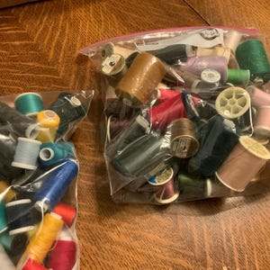 May include: Two clear plastic bags filled with spools of thread in various colors, including green, blue, yellow, red, and brown. The spools are of different sizes and materials, some with wooden cores. The bags are on a wooden surface.